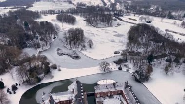 Karlı tarlalar, donmuş havuzlar, resmi bahçeler ve sakin beyaz bir manzarada yoğun ormanlık ile çevrili, kuleleri ve hendeği olan kırmızı tuğladan bir kalenin havadan görünüşü..