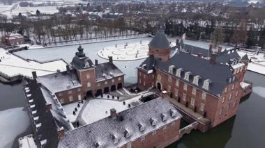 Dikenli çatıları, süslü kuleleri, karlı avlusu, hendeği ve çevresi orman ve evlerle çevrili donmuş simetrik bahçesi olan kırmızı tuğladan bir şatonun kış manzarası..
