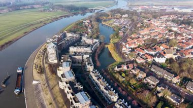 Çağdaş binalar, buhar yayan yüksek eğimli çatı kulesi, kenetlenmiş tekneler ve çevreleyen yeşil alanlar temiz bir yarı şehir manzarası oluşturan modern bir nehir kenarı bölgesi..