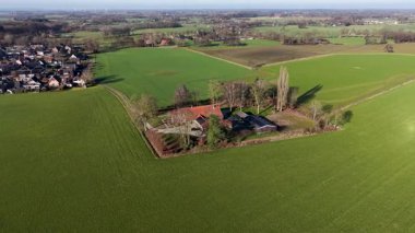 Geniş bir hava aracı görüntüsü geleneksel kırmızı çatılı bir çiftlik evinin canlı yeşil tarım arazileri ve sakin bir kır yolu ile çevrili ağaçların karesine yerleştirilmiş..