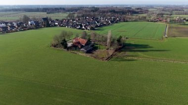 Geniş bir hava aracı görüntüsü geleneksel kırmızı çatılı bir çiftlik evinin canlı yeşil tarım arazileri ve sakin bir kır yolu ile çevrili ağaçların karesine yerleştirilmiş..