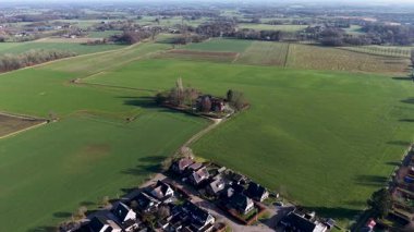 Geniş bir hava manzarası, açık bir gökyüzünün altında yemyeşil tarlalarla çevrili, kırmızı çatılı tenha bir çiftlik evini ve yakındaki bir köy kümesini gösteriyor..