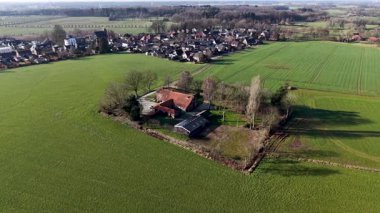 Geniş bir hava aracı görüntüsü geleneksel kırmızı çatılı bir çiftlik evinin canlı yeşil tarım arazileri ve sakin bir kır yolu ile çevrili ağaçların karesine yerleştirilmiş..