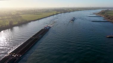 Aerial view of a long industrial barge transporting bulk cargo down a wide river at sunset. Wind turbines and misty green fields line the horizon under a soft golden glow.