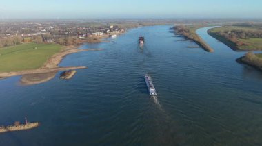 Endüstriyel mavnalardan oluşan geniş bir hava manzarası geniş mavi bir nehirden modern bir kablo destekli köprüye doğru ilerliyor. Sahnede yemyeşil bankalar ve temiz, parlak bir gökyüzü altında tarihi bir kasaba yer alıyor..