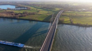 Aerial view of a modern cable stayed bridge over a wide river. Industrial vessels navigate the water between green banks under a hazy, sunlit sky in a peaceful European landscape.