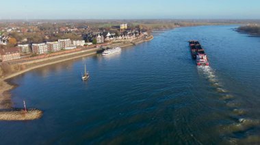Endüstriyel bir mavnanın, Avrupa 'nın pitoresk bir kasabasının yanındaki geniş mavi bir nehirde yüzüşü. Sahnede modern ve geleneksel mimari berrak, parlak bir gökyüzü altında yer alıyor..