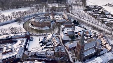 Karla kaplı bir kasabanın içine yerleştirilmiş tarihi bir şatonun yüksek açılı kış hava manzarası. Sahnede dondurulmuş ağaçlar, bir kilise kulesi ve yumuşak gün ışığı altında geleneksel binalar yer alıyor..