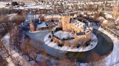 Kışın tarihi bir şatonun havadaki altın saat manzarası. Kar tuğla kalenin ve köyün etrafını sararken, uzun gölgeler kısmen donmuş dairesel hendek boyunca uzanıyor..