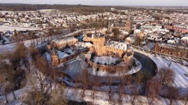 Kışın tarihi bir şatonun havadaki altın saat manzarası. Kar tuğla kalenin ve köyün etrafını sararken, uzun gölgeler kısmen donmuş dairesel hendek boyunca uzanıyor..