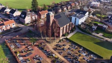 Bir Avrupa köy merkezinin bilgilendirici hava görüntüsü, tarihi tuğla bir kilise ve güneş panelleri olan modern yerleşim evleriyle çevrili komşu bir mezarlık içeriyor..