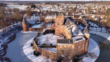 Güneş ışığı, karlı bir Avrupa manzarasında tarihi bir şatoyu aydınlatır. Tuğla kalede dairesel donmuş bir hendek, taş kuleler ve berrak bir kış gökyüzünün altında donmuş ağaçlar yer alıyor..