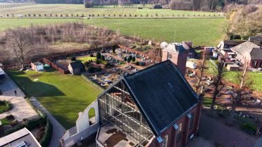 Çarpıcı modern cam kaplamalı tarihi tuğla bir kilisenin yer aldığı bir Avrupa köyünün havadan görünüşü. Sahnede yerel bir mezarlık ve güneş panelleri olan parlak güneş ışığı altındaki evler yer alıyor..