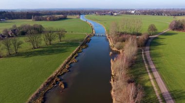 Yeşil tarlalar boyunca uzanan kanalı olan kırsal bir arazinin havadan görünüşü, içinden geçen iki küçük yol köprüsü, ve açık tarım arazileri arasında ek binalar bulunan yakın bir ev..
