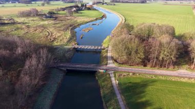 Bereketli yeşil tarlalar ve ormanlık alanlarda dolanan, yüksek açılı mavi bir kanalın insansız hava aracı görüntüsü, iki köprü, bir bisiklet yolu ve yumuşak güneş ışığı altında küçük bir köy..