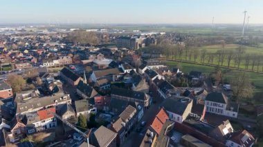 Geleneksel tuğla mimarisi, merkezi bir kilise ve dar sokakların yer aldığı bir Avrupa kasabasının havadan görünüşü. Arka plan, günbatımında bir sanayi bölgesi, yeşil alanlar ve rüzgar türbinlerini gösteriyor..