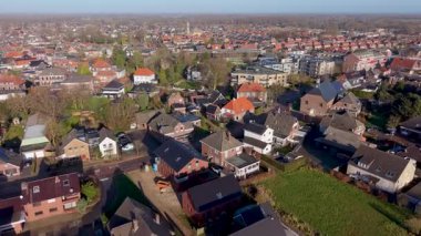Geleneksel kırmızı kiremitli çatıları, modern tuğlalı evleri, yemyeşil bahçeleri ve temiz güneş ışığı altında uzak bir kilise kulesi olan pitoresk bir Avrupa kasabasını gösteren yüksek açılı hava aracı görüntüsü..