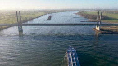 Bir kargo gemisi ve tankerin kablo altında büyük bir nehirde yüzüşü gündoğumunda köprüde kaldı. Avrupa 'da manzaralı bir nehir manzarasında sanayi deniz taşımacılığı ve lojistik.