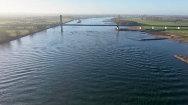 Modern bir kablonun hava görüntüsü gündoğumunda geniş bir nehri geçerek köprü olarak kaldı. Yeşil çayırlar, nehir yosunları ve sisli mavi gökyüzünün altında uzak rüzgar türbinleri..