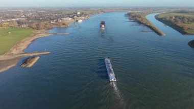 Geniş bir nehirde yol alan iki büyük kargo gemisinin havadan görüntüsü. Çimenli bankalar, küçük yarımadalar ve çevresi yeşil tarlalarla çevrili bir kasaba ve sakin bir kırsal alanda dağınık evler..
