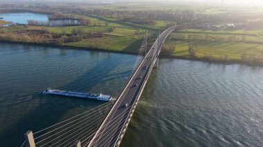Kablo görüntüsü geniş bir nehir üzerinde köprü vazifesi görürken, aşağıda yeşil tarlalar, kırsal evler, rüzgar türbinleri ve sakin kırsal manzaralarla çevrili büyük bir kargo gemisi vardı..