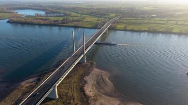 Kablo görüntüsü geniş bir nehir üzerinde köprü vazifesi görürken, aşağıda yeşil tarlalar, kırsal evler, rüzgar türbinleri ve sakin kırsal manzaralarla çevrili büyük bir kargo gemisi vardı..