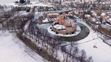 Karlı kış mevsiminde donmuş bir hendekle çevrili tarihi bir tuğla şatonun havadan görünüşü yapraksız ağaçlar, açık alanlar ve yüksek kilise kulesi olan yakın bir kasaba..