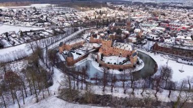 Karlı bir kasabayla çevrili tarihi bir tuğla şatonun havadan görünüşü. Donmuş hendekler, yapraksız ağaçlar, yoğun binalar ve güzel bir kış manzarası yaratan uzun bir kilise kulesi..