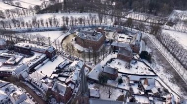 Karlı bir kasabayla çevrili tarihi bir tuğla şatonun hava manzarası. Donmuş hendekler, yapraksız ağaçlar, açık alanlar ve güzel bir kış manzarası yaratan uzun bir kilise kulesi..