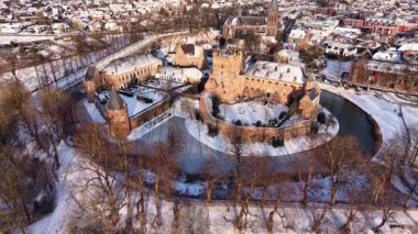 Kışın tarihi bir şatonun havadaki altın saat manzarası. Kar tuğla kalenin ve köyün etrafını sararken, uzun gölgeler kısmen donmuş dairesel hendek boyunca uzanıyor..