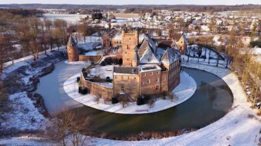 Kışın tarihi bir şatonun havadaki altın saat manzarası. Kar tuğla kalenin ve köyün etrafını sararken, uzun gölgeler kısmen donmuş dairesel hendek boyunca uzanıyor..