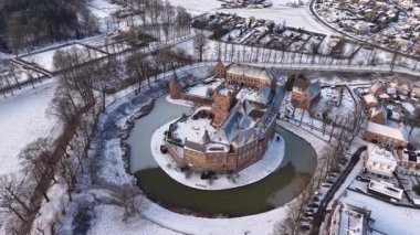Kışın tarihi bir şatonun havadaki altın saat manzarası. Kar tuğla kalenin ve köyün etrafını sararken, uzun gölgeler kısmen donmuş dairesel hendek boyunca uzanıyor..