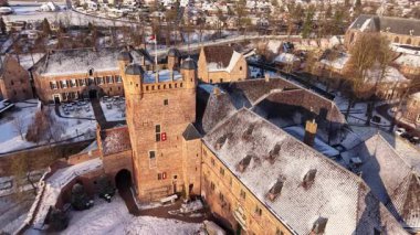 Güneş ışığı, karlı bir Avrupa manzarasında tarihi bir şatoyu aydınlatır. Tuğla kalede dairesel donmuş bir hendek, taş kuleler ve berrak bir kış gökyüzünün altında donmuş ağaçlar yer alıyor..