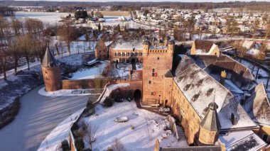 Güneş ışığı, karlı bir Avrupa manzarasında tarihi bir şatoyu aydınlatır. Tuğla kalede dairesel donmuş bir hendek, taş kuleler ve berrak bir kış gökyüzünün altında donmuş ağaçlar yer alıyor..