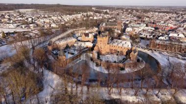 Kışın tarihi bir şatonun havadaki altın saat manzarası. Kar tuğla kalenin ve köyün etrafını sararken, uzun gölgeler kısmen donmuş dairesel hendek boyunca uzanıyor..