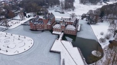 Yapraksız ağaçlar ve açık karlı tarlalarla çevrili kar kaplı resmi bir bahçenin yanında, kuleleri ve kısmen donmuş hendeği olan kırmızı tuğladan bir kalenin havadan görünüşü..