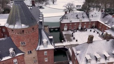 Kızıl tuğlalı bir kalenin havadan görünüşü, sarp kayalık çatılar, kısmen donmuş bir hendek, bir köprü girişi, ve tarihi kompleksi çevreleyen karla kaplı ağaçlar ve tarlalar..