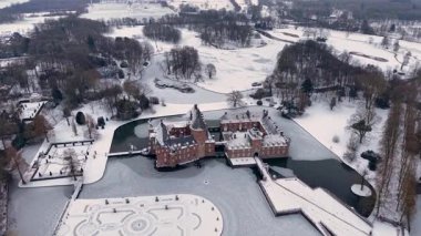 Yapraksız ağaçlar ve açık karlı tarlalarla çevrili kar kaplı resmi bir bahçenin yanında, kuleleri ve kısmen donmuş hendeği olan kırmızı tuğladan bir kalenin havadan görünüşü..