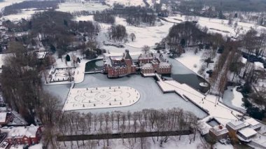 Yapraksız ağaçlar ve açık karlı tarlalarla çevrili kar kaplı resmi bir bahçenin yanında, kuleleri ve kısmen donmuş hendeği olan kırmızı tuğladan bir kalenin havadan görünüşü..