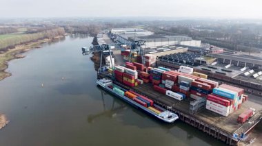 Endüstriyel bir liman terminalindeki nakliye konteynırlarını yükleyen nehir mavnasının hava görüntüsü. Açık bir gökyüzü altında bir vinç, lojistik depoları ve kırsal alan var..