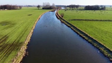 Yeşil tarlalar boyunca uzanan kanalı olan kırsal bir arazinin havadan görünüşü, içinden geçen iki küçük yol köprüsü, ve açık tarım arazileri arasında ek binalar bulunan yakın bir ev..