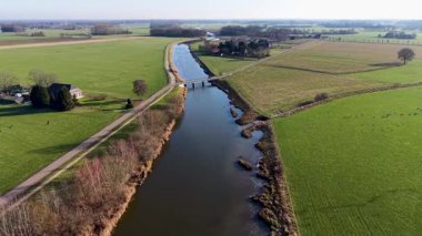 Yeşil tarlalar boyunca uzanan kanalı olan kırsal bir arazinin havadan görünüşü, içinden geçen iki küçük yol köprüsü, ve açık tarım arazileri arasında ek binalar bulunan yakın bir ev..