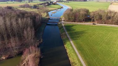 Yeşil tarlalar boyunca uzanan kanalı olan kırsal bir arazinin havadan görünüşü, içinden geçen iki küçük yol köprüsü, ve açık tarım arazileri arasında ek binalar bulunan yakın bir ev..