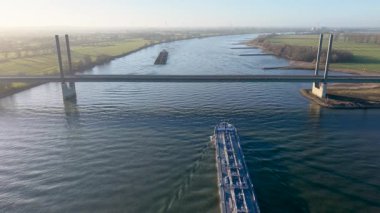 Geniş bir nehirde, kablo destekli modern bir köprünün altından geçen bir kargo gemisinin hava görüntüsü. Yeşil bankaları, rüzgar türbinleri ve suya yansıyan yumuşak sabah güneşiyle Avrupa manzarası.