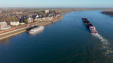 Bir kargo mavnası ve tarihi bir Avrupa kasabasının yanındaki geniş bir nehirdeki yolcu gemisinin havadan görüntüsü. Manzaralı nehir kenarı mimarisi, taş setler, ve parlak güneşli bir gökyüzünün altında berrak mavi su..