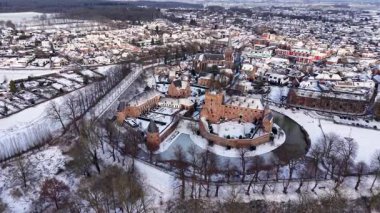 Kışın donmuş hendeği olan bir ortaçağ şatosunun çarpıcı hava manzarası. Soğuk kış gökyüzü altında bir kilise ve geleneksel evler ile karla kaplı Avrupa şehir manzarası. Tarihsel kış manzarası.
