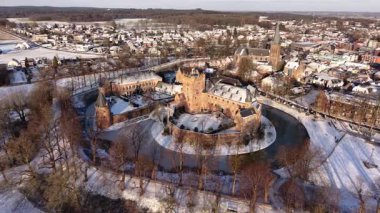 Kışın donmuş hendeği olan bir ortaçağ şatosunun çarpıcı hava manzarası. Soğuk kış gökyüzü altında bir kilise ve geleneksel evler ile karla kaplı Avrupa şehir manzarası. Tarihsel kış manzarası.