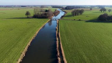 Yeşil tarlalar boyunca uzanan kanalı olan kırsal bir arazinin havadan görünüşü, içinden geçen iki küçük yol köprüsü, ve açık tarım arazileri arasında ek binalar bulunan yakın bir ev..