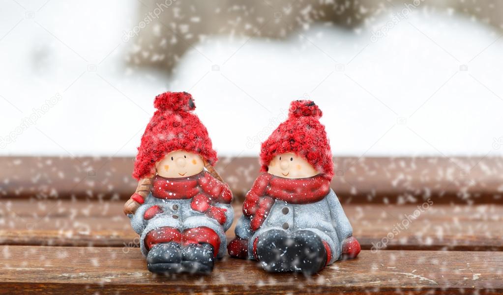 two dolls sitting on bench at winter day. snowing