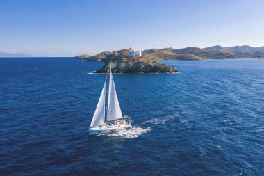 Yelkenli. Beyaz yelkenli yelkenli, dalgalı deniz arka planı, pelerinli deniz feneri. Yunanistan, Kea Tzia adası. Ege Denizi 'nde yaz tatili. Hava aracı görünümü